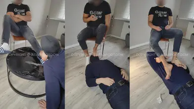 Un dominant arabe scrolle son téléphone d'une main pendant qu'un homme cagoulé rampe à ses pieds nus
